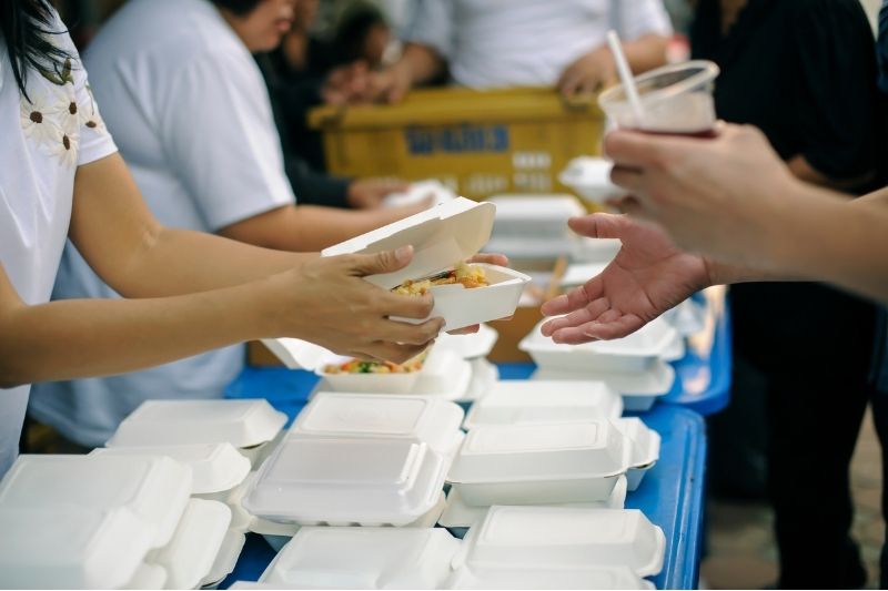 Volunteers handing out meals to people standing in line on a hot afternoon.